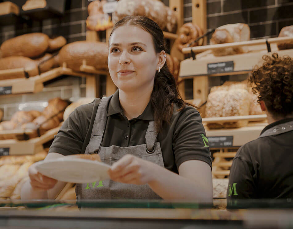 Verkäuferin-Azubi lächelt in der Bäckerei Meyer mit vielen Broten im Hintergrund