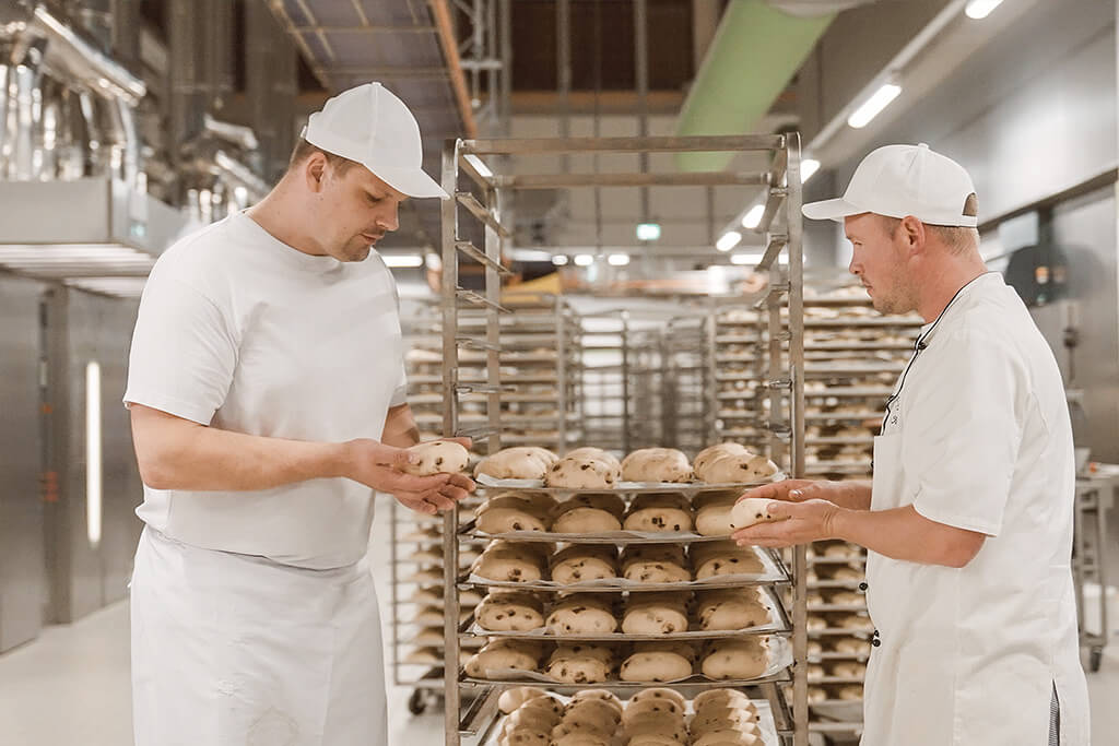 Zwei Bäcker stehen in der Backstube vor einem Wagen mit ungebackenen Brötchen und prüfen die Qualität des Teiges.