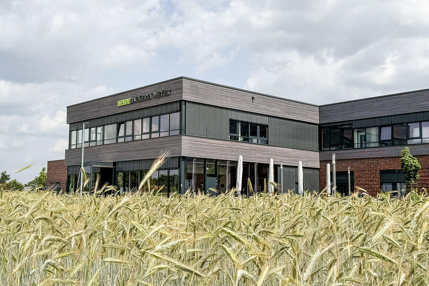Großes Produktionsgebäuse mit Holz- Backstein und Glasfronten. Davor ein Roggenfeld.